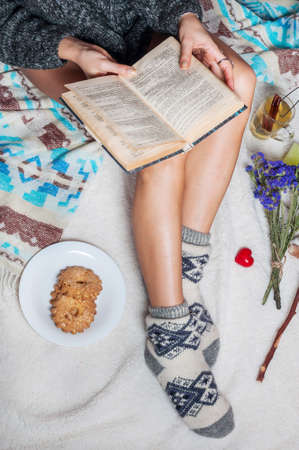 Long female feet in warm woolen socks with cup of tea and book.