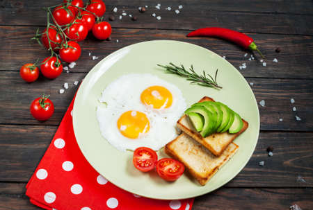 Pan of fried eggs and cherry-tomatoes with bread, avocado on dark wood table.