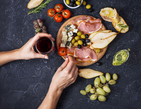 Woman hands hold a glass of wine. Appetizer, italian antipasto, ham, olives, cheese, bread, grapes, pear on dark stone background. Top viewの写真素材
