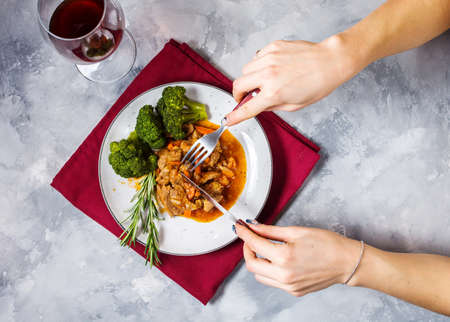 Beef goulash with vegetables and broccoli on rustic concrete background, women's hands hold fork and knife. Top viewの写真素材