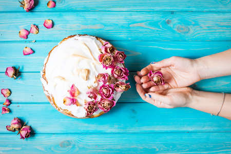 Woman's hands hold the dry rose. The white cream cake decorated dry rose on blue wood background. Top viewの写真素材