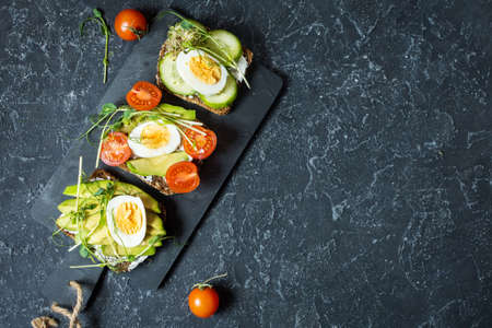 Tasty sandwiches with egg, avocado and vegetables on stone black background. Copy space