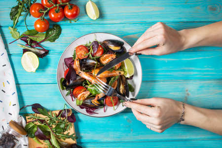 Fresh seafood salad, mussels, shrimp, fresh vegetables and herbs on blue wooden table. Female hands...