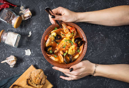 French fish soup Bouillabaisse with seafood, salmon fillet, shrimp, mussels on black stone background. Female hands hold spoon. Top viewの写真素材