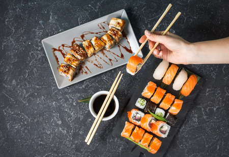 Female hand hold sushi roll with chopsticks on black stone background. Top view