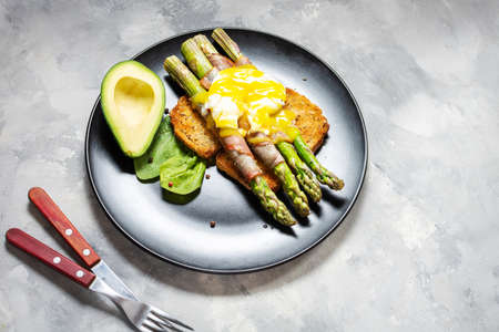 Grilled green asparagus wrapped with bacon, benedict poached egg and avocado on toasts for breakfast