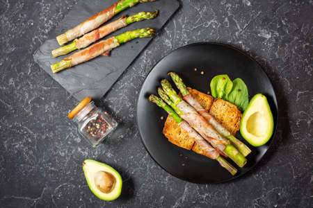 Grilled green asparagus wrapped with bacon and avocado on toastsの写真素材