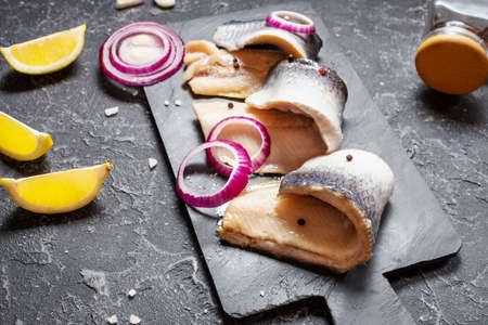 Herring fillet with salt, herbs, onion and lemon on black plate on stone background. Healthy food