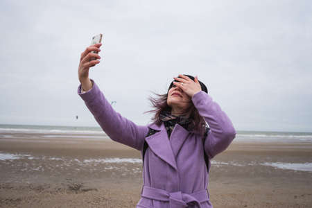 Cute attractive woman in stylish coat smiles and takes selfie outside on the seafront of Dunkerk in the north of France on a cloudy spring day. Having fun.の写真素材