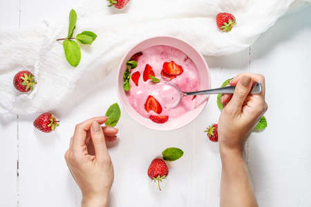 Strawberry ice cream in a pink bowl with strawberries on a concrete background. Female hand hold a spoon. Top viewの写真素材