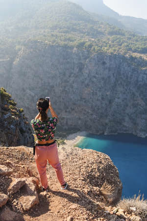 Young woman taking pictures on smartphone at the Butterfly Valley from above, sitting on the rocks....