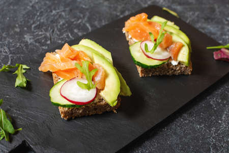 open sanwiches with dark rye bread, avocado, smoked salmon, cucumber and radish on black stone...