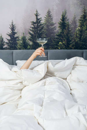 Woman is lying in bed raising hand holding glass filled with drink. Care free and irresponsible...