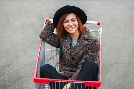 Cheerful stylish young girl in a coat and hat sitting in a shopping cart in a supermarket.の写真素材
