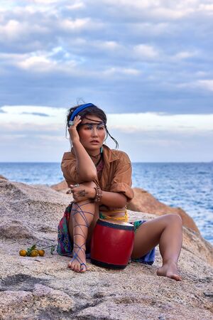 Young woman shaman in bright clothes and makeup, with drum, seating in the mountain, sea view. Ethnic fashion photoshoot. Boho style.の写真素材