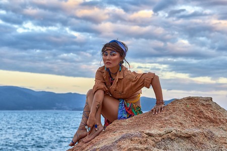 Asian Woman with bright fashion makeup, ethnic clothes in boho style, seating on rocky mountain near the sea. Beauty photoshoot in ethnic fashion style.の写真素材
