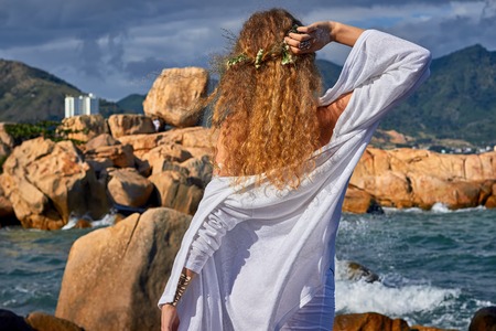 portrait of pretty sensual curly girl with flowers in her head, white dress, standing in a stone...