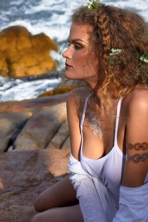 portrait of pretty sensual curly girl with flowers in her head, white dress, seating in a stone...