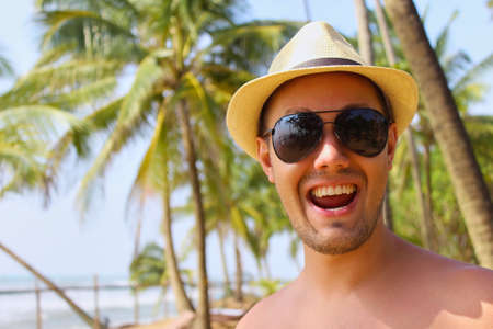Young man smilling on a tropical  summer beach, happy mood, wearing sunglasses, palm trees background. Travelling vacation photo.の写真素材