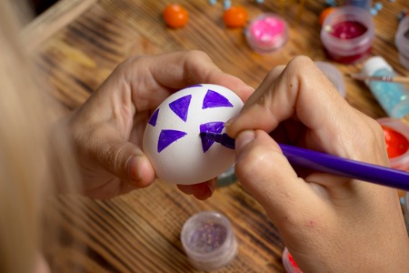 girl artist  paints easter egg with purple marker, process of creating holiday decorations, paint cans, glitters, candies on wooden tableの写真素材