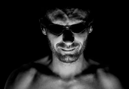 Portrait of unshaven adult caucasian man in swimming glasses. He smiles and looks straight to watcher. Black and white shot, low-key lighting. Isolated on black.の写真素材