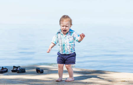 Cute baby boy is crying on the beach with a beautiful blue sea in the background. Portrait of an alone sad child on a beach in a sunny day.の写真素材