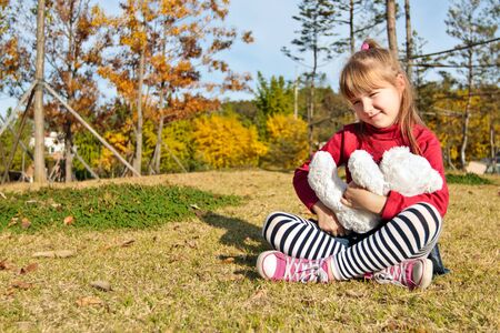 Little girl with white bear toy sitting on a grass in autumn parkの写真素材