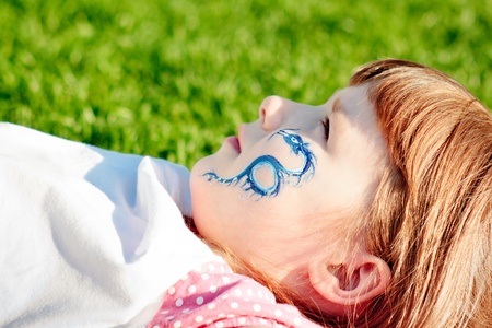 Portrait of a young girl with blue dragon on her face lying on a lawn