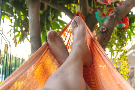 Close-up of bare feet resting in an orange hammock under a tree.の写真素材