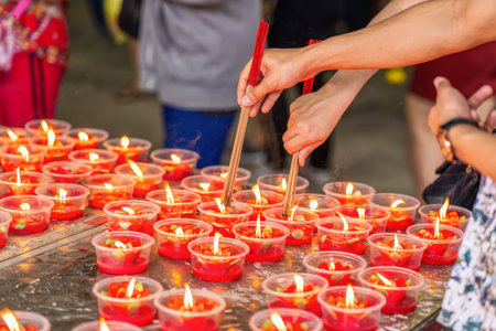 Hands holding incense sticks over rows of small red devotional candles, flames flickering brightly during a temple prayer ritual.の写真素材