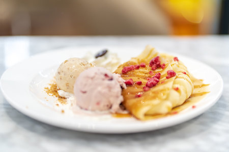 Soft focus dessert plate featuring folded crepes drizzled with syrup and topped with raspberries, served with two scoops of ice creamの写真素材