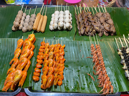 A variety of Thai street food skewers including meatballs, sausages, and small shrimps displayed on fresh green banana leaves.の写真素材