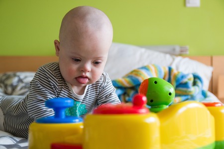 Cute baby boy with Down syndrome playing with toy on the bed in home bedroomの写真素材