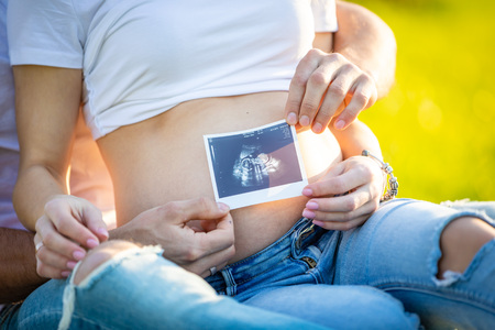 Happy couple expecting baby, pregnant young woman with husband sitting on green grass and holding...