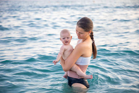 Beautiful young woman with her daughter little baby girl swimming in the sea at sunset time. Woman with daughter relaxing in front of seaの写真素材