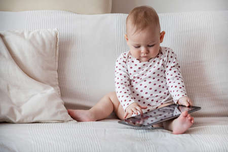 Baby girl sitting on white sofa and holding grey tabletの写真素材