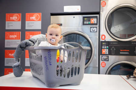 Prague, Czech Republic - 16.12.2020: Baby girl in the plastic basket waiting in laundry room with Speed Queen self service coin-operated washing machine and dry cloth laundry, Prague, Czech Republicのeditorial素材