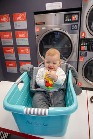 Prague, Czech Republic - 16.12.2020: Baby girl in the plastic basket waiting in laundry room with Speed Queen self service coin-operated washing machine and dry cloth laundry, Prague, Czech Republicのeditorial素材