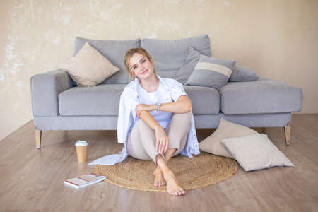 Beautiful sensual young woman with blonde hair wearing a white shirt reading book and drinking coffee on the sofa in modern homeの写真素材