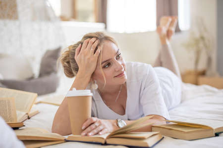 Beautiful sensual young woman with blonde hair wearing a white shirt reading books and drinking...