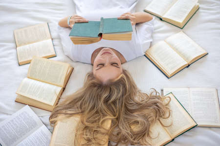 Beautiful sensual young woman with blonde hair wearing a white shirt reading books on the bed in modern homeの写真素材