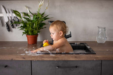 Cute happy baby girl with playing with water and foam in a kitchen sink at homeの写真素材