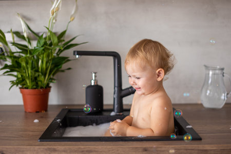 Cute happy baby girl with playing with water and foam in a kitchen sink at homeの写真素材