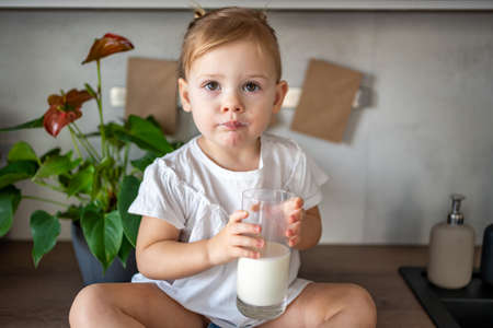 Happy baby girl sitting at the table in the kitchen and drinking milkの写真素材