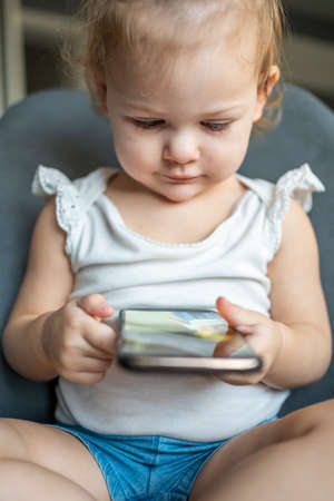 Smiling little girl is sitting on armchair and using smart phoneの写真素材