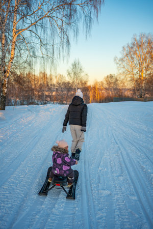 Young woman pulling plastic sled with little child across snowy landscape. Winter nature. Family.の写真素材