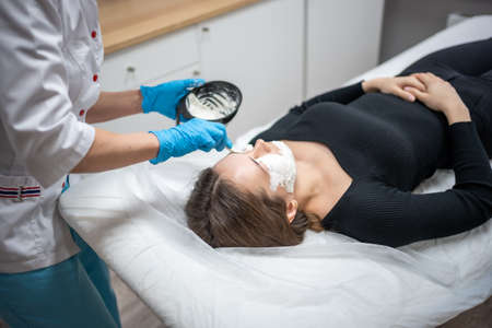 Doctor cosmetologist or dermatologist making face mask in cosmetology salon. Professional Beautician applying face mask on caucasian woman face lying on bed in bathrobe.の写真素材