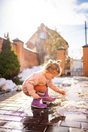 Girl plays in a puddle with wooden stick in spring day at sunlightの写真素材