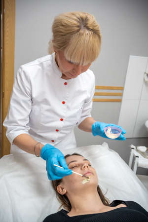 Doctor cosmetologist or dermatologist making face mask in cosmetology salon. Professional Beautician applying face mask on caucasian woman face lying on bed in bathrobe.の写真素材