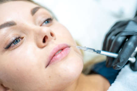 Close-up of woman face and hand in surgical glove holding syringe near her lips, ready to receive...